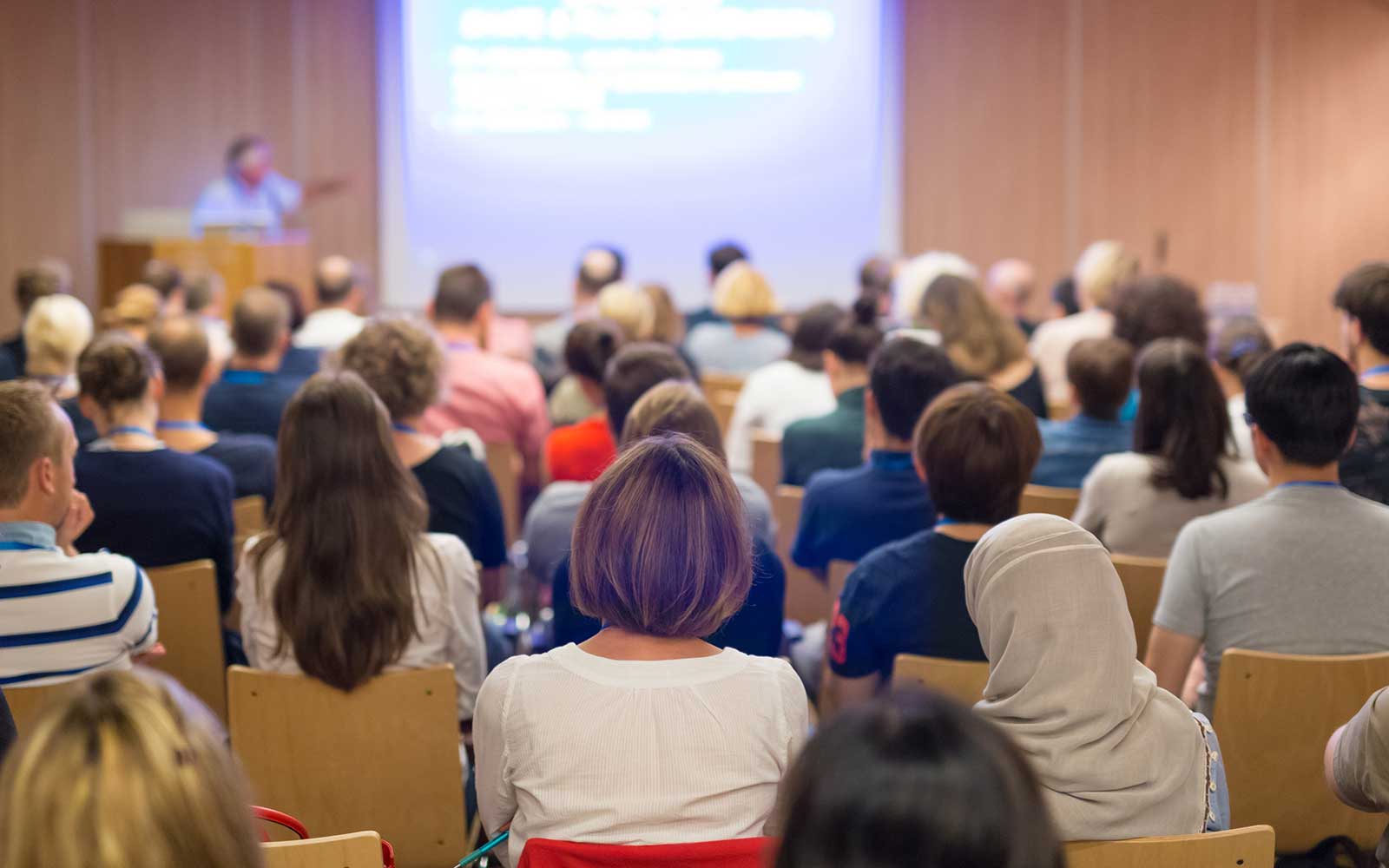 View from the rear of a seated group in a large room where a presentation with slides is being delivered by a speaker at a podium. 