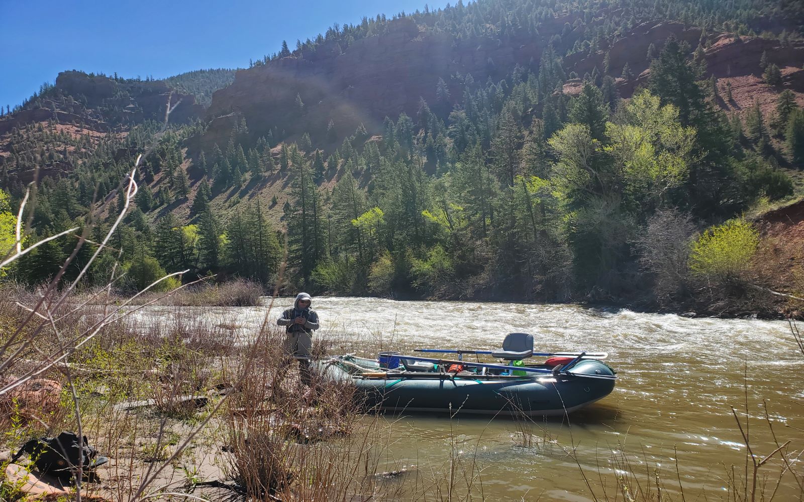 A man stands by a raft in a river flowing through tree-covered hills