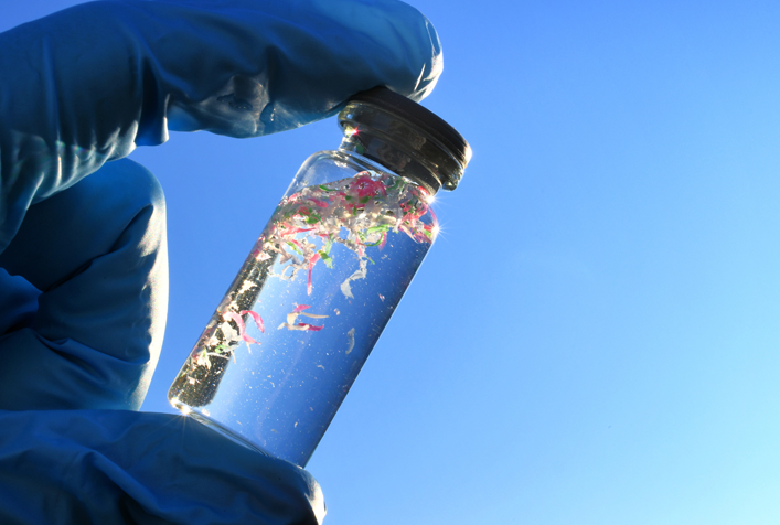 Close up image of a gloved hand holding a small glass vile containing micro plastics