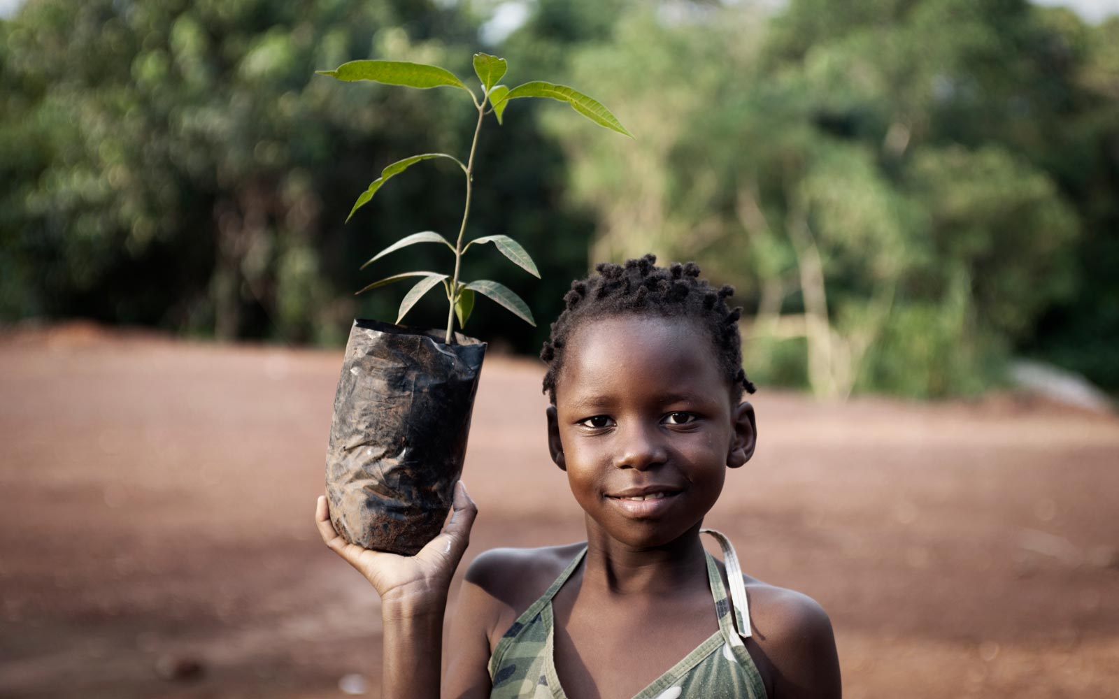 African child in a rural African setting, holding a bagged plant ready for planting.