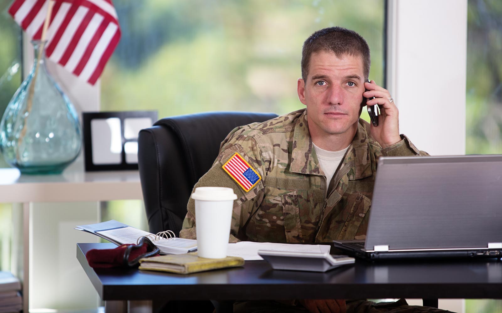 Male veteran on a phone at a work desk with an American flag in the background.