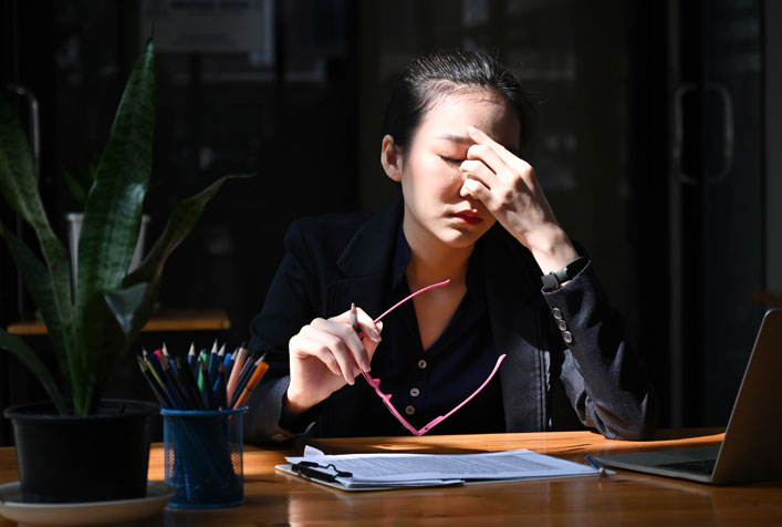 Photo of a person sitting at a table with paperwork, holding her glasses in one hand while the other is pinching the bridge of nose as if they have a headache